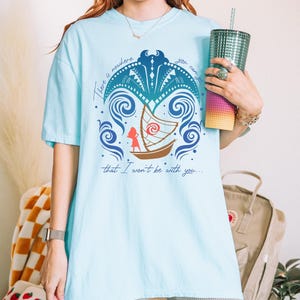 May include: Light blue t-shirt with a graphic design featuring a boat, waves, and the text "There is nowhere you can that I won't be with you...". The design is in shades of blue, brown, and red.