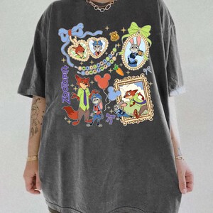 May include: Dark gray Zootopia graphic t-shirt featuring cartoon characters Nick Wilde and Judy Hopps. The design includes framed portraits, balloons, and the word "Zootopia" in a stylized font. The shirt has short sleeves and a relaxed fit.