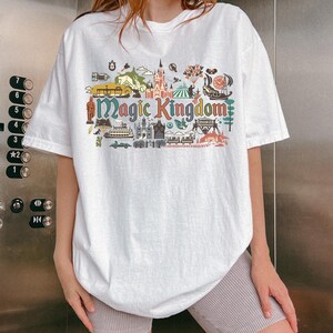 May include: White t-shirt with a colorful graphic design featuring the words "Magic Kingdom" and various illustrations of amusement park attractions. The shirt has short sleeves and a relaxed fit.