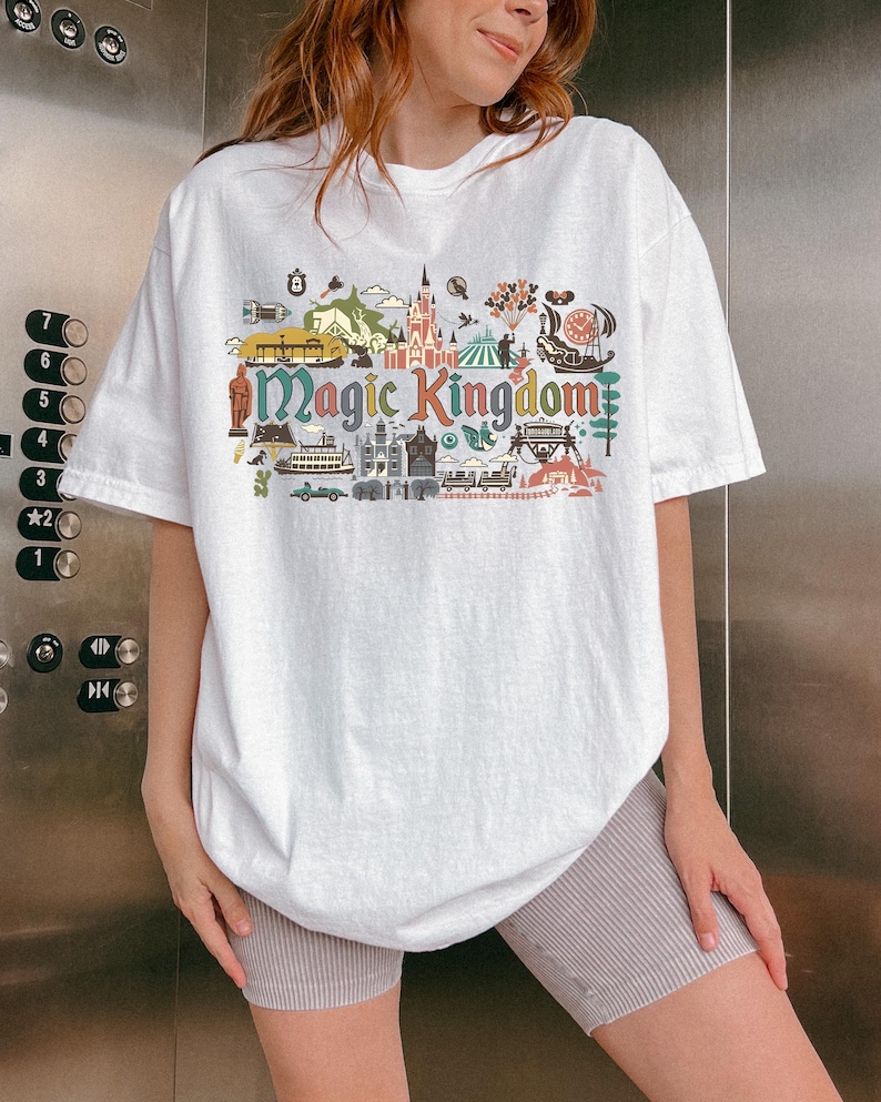May include: White t-shirt with a colorful graphic design featuring the words "Magic Kingdom" and various illustrations of amusement park attractions. The shirt has short sleeves and a relaxed fit.