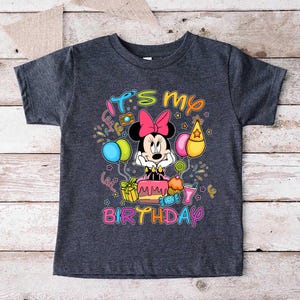 May include: Dark gray t-shirt featuring a colorful graphic of Minnie Mouse with a pink bow, a birthday cake, and balloons. The text "It's my birthday" is written in a playful font. A fun, festive design for a child's birthday.