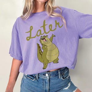 May include: A lilac-colored t-shirt featuring a cartoon alligator with the word "Later" in a cursive font. The alligator is green and tan, with its arms raised. The shirt is made of a soft material.
