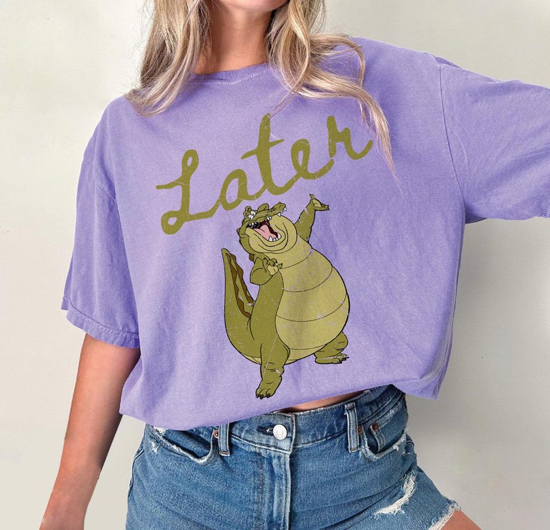 May include: A lilac-colored t-shirt featuring a cartoon alligator with the word "Later" in a cursive font. The alligator is green and tan, with its arms raised. The shirt is made of a soft material.