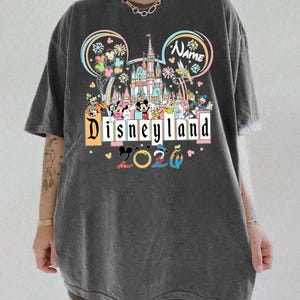 Op de afbeelding: Donkergrijs T-shirt met een Disneyland-ontwerp met Mickey Mouse, Minnie Mouse en andere Disney-personages. Het ontwerp bevat een kasteel, het woord "Disneyland" en het jaar "2020". Het shirt heeft korte mouwen en een relaxte pasvorm.