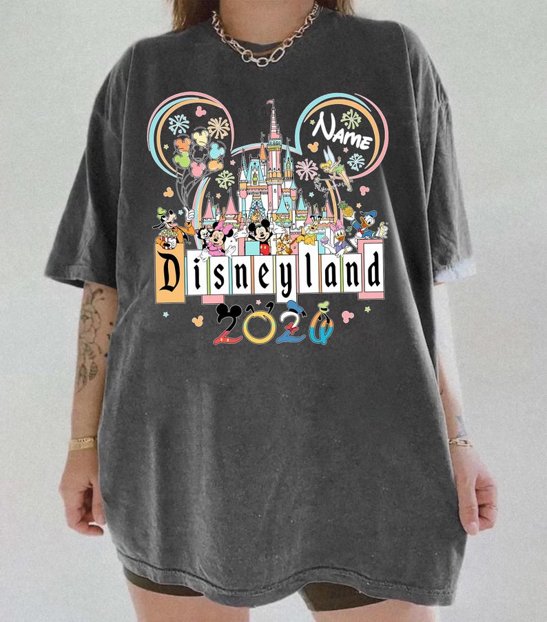 Op de afbeelding: Donkergrijs T-shirt met een Disneyland-ontwerp met Mickey Mouse, Minnie Mouse en andere Disney-personages. Het ontwerp bevat een kasteel, het woord "Disneyland" en het jaar "2020". Het shirt heeft korte mouwen en een relaxte pasvorm.