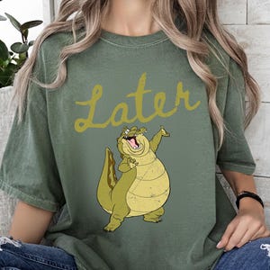 May include: Olive green t-shirt with the word "Later" in a yellow script font. A cartoon alligator with a wide grin and raised arm is printed below the text. The alligator is yellow and green.