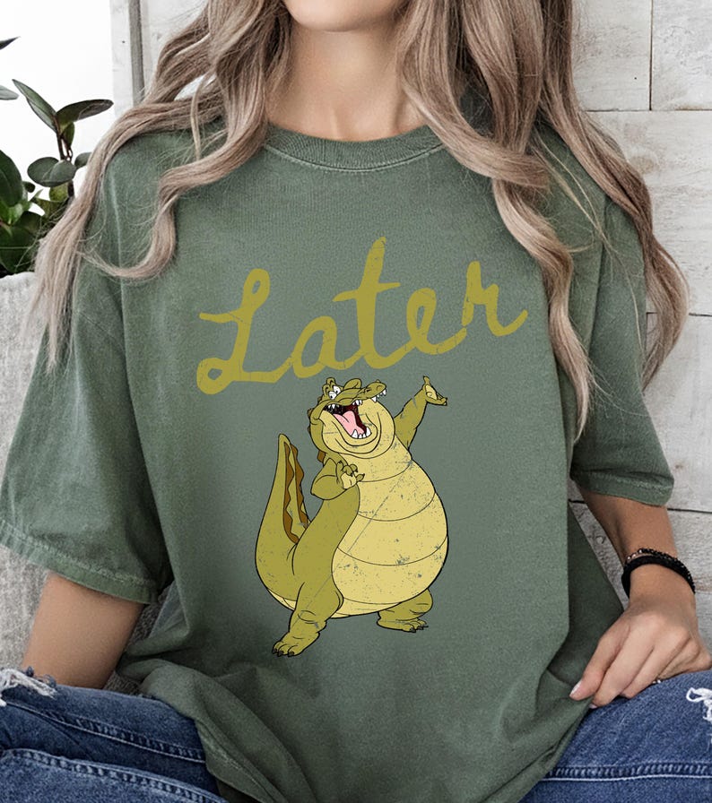 May include: Olive green t-shirt with the word "Later" in a yellow script font. A cartoon alligator with a wide grin and raised arm is printed below the text. The alligator is yellow and green.