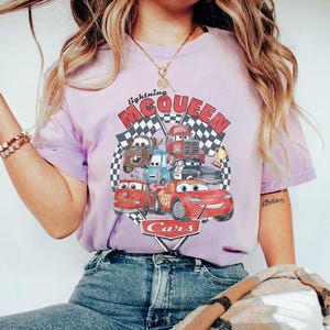Puede incluir: Camiseta lila claro con un dise&ntilde;o de personajes de la pel&iacute;cula Cars, incluyendo a Rayo McQueen. El dise&ntilde;o incluye el texto "Lightning McQueen" y "Cars" sobre un fondo de bandera a cuadros.