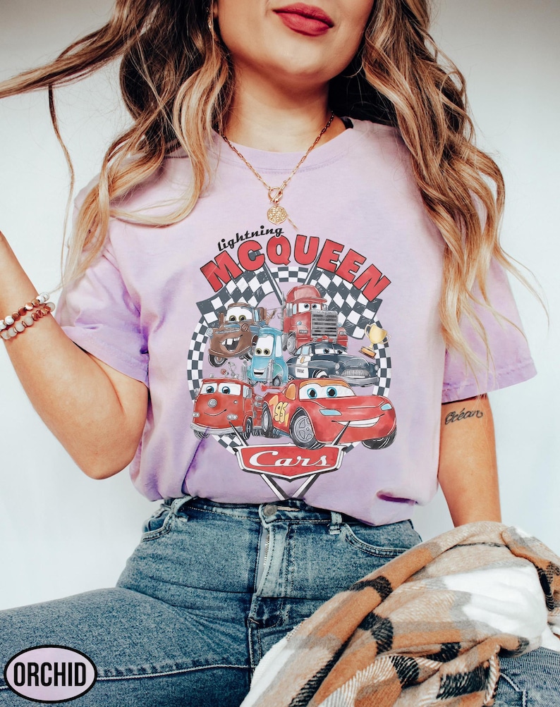 Puede incluir: Camiseta lila claro con un dise&ntilde;o de personajes de la pel&iacute;cula Cars, incluyendo a Rayo McQueen. El dise&ntilde;o incluye el texto "Lightning McQueen" y "Cars" sobre un fondo de bandera a cuadros.