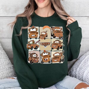 Może przedstawiać: Ciemnozielona bluza z siatką z kresk&oacute;wkowymi obrazami Złomka z filmu Auta. Obrazy są w odcieniach brązu i beżu, ze słowem "Cars" w beżowym prostokącie.