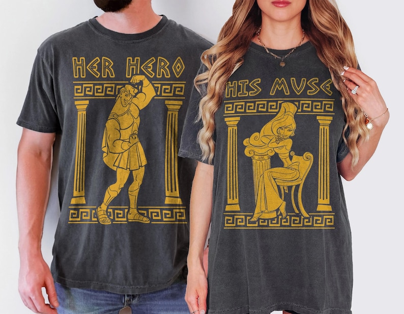 May include: Two dark gray t-shirts with gold-colored graphics. One shirt says "HER HERO" with an illustration of a muscular figure. The other says "HIS MUSE" with a seated woman. Both shirts have a vintage, distressed look.