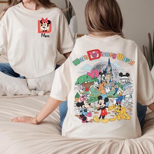 May include: Cream-colored t-shirt featuring a Minnie Mouse graphic with the word "Mom" on the front. The back of the shirt displays a colorful Walt Disney World design with various cartoon characters, including Mickey Mouse and Donald Duck.