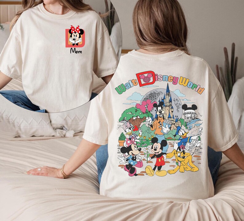 May include: Cream-colored t-shirt featuring a Minnie Mouse graphic with the word "Mom" on the front. The back of the shirt displays a colorful Walt Disney World design with various cartoon characters, including Mickey Mouse and Donald Duck.