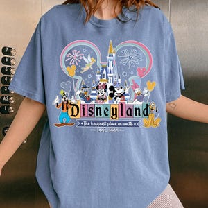 Może przedstawiać: Jasnoniebieska koszulka z kolorową grafiką Disneylandu. Projekt zawiera Myszkę Miki i Minnie, Kaczora Donalda, Goofy'ego i inne postacie Disneya. Tekst brzmi "Disneyland" i "The happiest place on earth. EST. 1955."