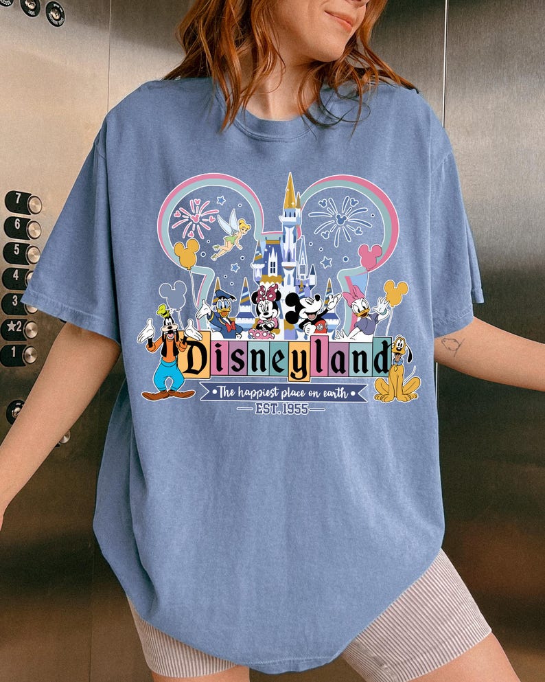 Może przedstawiać: Jasnoniebieska koszulka z kolorową grafiką Disneylandu. Projekt zawiera Myszkę Miki i Minnie, Kaczora Donalda, Goofy'ego i inne postacie Disneya. Tekst brzmi "Disneyland" i "The happiest place on earth. EST. 1955."
