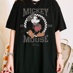 May include: Black t-shirt featuring a distressed graphic of Mickey Mouse. The design includes the words "Mickey Mouse" and "1928" in a vintage font, with Mickey in his classic pose. The Disney logo is at the bottom.