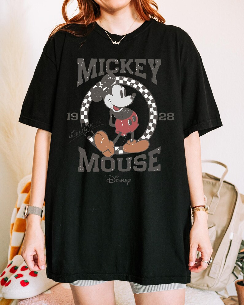 May include: Black t-shirt featuring a distressed graphic of Mickey Mouse. The design includes the words "Mickey Mouse" and "1928" in a vintage font, with Mickey in his classic pose. The Disney logo is at the bottom.