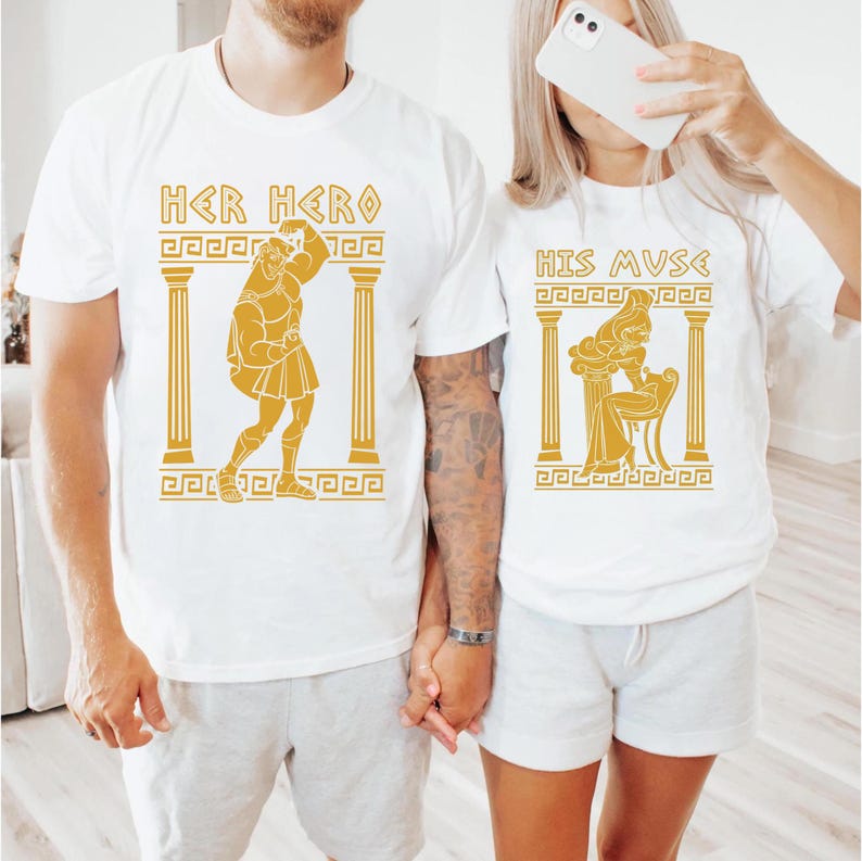 May include: White t-shirts with gold Greek-inspired designs. One shirt reads "HER HERO" with a graphic of a muscular figure. The other shirt says "HIS MUSE" with a seated female figure. The couple is holding hands.