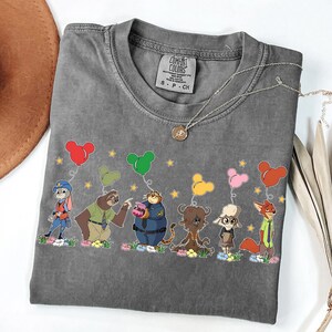 以下が含まれることがあります： ミッキーマウスの形をした風船を持っている漫画の動物のカラフルなグラフィックが特徴のグレーのコンフォートカラーズTシャツ。 シャツはクルーネックと半袖です。 ペンダント付きのシルバーのネックレスがシャツの上にかけられています。