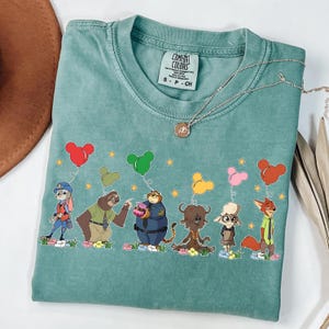 以下が含まれることがあります： ズートピアの漫画のキャラクターがミッキーマウスの形をした風船を持っている、ターコイズ色のComfort Colors Tシャツ。 シャツはクルーネックと半袖です。 ペンダント付きのシルバーのネックレスがシャツにかけられています。