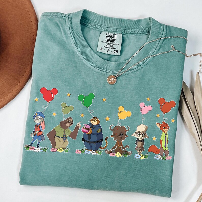 以下が含まれることがあります： ズートピアの漫画のキャラクターがミッキーマウスの形をした風船を持っている、ターコイズ色のComfort Colors Tシャツ。 シャツはクルーネックと半袖です。 ペンダント付きのシルバーのネックレスがシャツにかけられています。