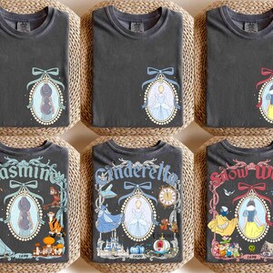 May include: Dark gray t-shirts with various princess designs. Each shirt features a framed portrait of a princess, with the names Jasmine, Cinderella, and Snow White. The shirts are folded on woven mats.