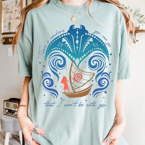 May include: A light green t-shirt featuring a boat with a character illustration and the text "There is nowhere you can go that I won't be with you...". The design includes blue waves and decorative elements.