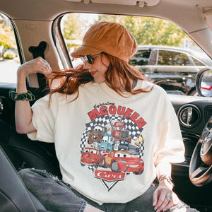 Puede incluir: Camiseta de color crema con un gr&aacute;fico de Rayo McQueen y otros personajes de la pel&iacute;cula Cars. El dise&ntilde;o incluye el texto "lightning McQueen" en rojo y negro, con un fondo de bandera a cuadros. La camiseta la lleva una persona.