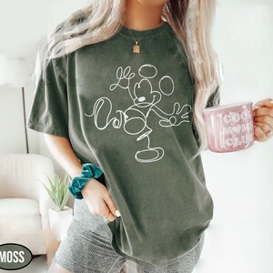 May include: Moss green t-shirt featuring a white line drawing of Mickey Mouse. The shirt has a relaxed fit. A pink mug with the words "Cool Mom Club" is held in the hand. The word "MOSS" is in a black oval.