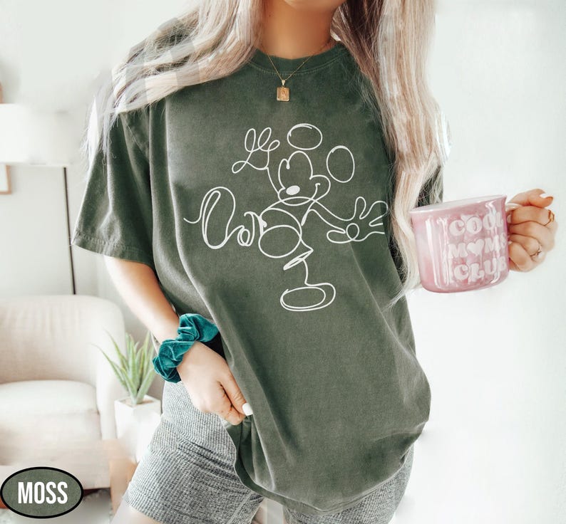 May include: Moss green t-shirt featuring a white line drawing of Mickey Mouse. The shirt has a relaxed fit. A pink mug with the words "Cool Mom Club" is held in the hand. The word "MOSS" is in a black oval.