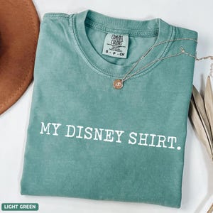 May include: Light green Comfort Colors t-shirt with the text "MY DISNEY SHIRT." A silver necklace with a pendant is draped across the shirt. The shirt is folded and has a tag that says "S - P - CH."