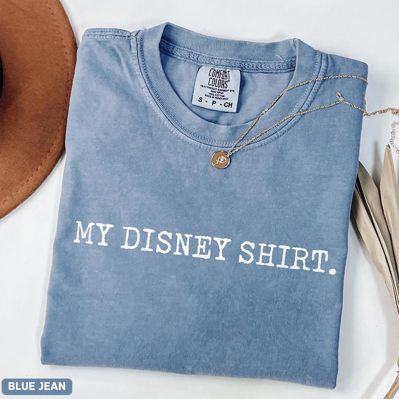 May include: A blue jean Comfort Colors t-shirt with the text "MY DISNEY SHIRT." A gold necklace with a pendant rests on the shirt. A brown hat and dried plants are in the background.