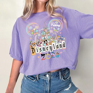 Op de afbeelding: Een lila T-shirt met een Disneyland-afbeelding. Het ontwerp bevat een kasteel, Mickey en Minnie Mouse en andere Disney-personages. De tekst "Disneyland 2020" is ook zichtbaar.