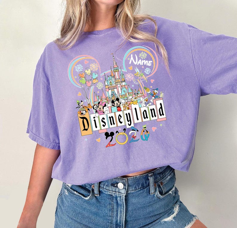 Op de afbeelding: Een lila T-shirt met een Disneyland-afbeelding. Het ontwerp bevat een kasteel, Mickey en Minnie Mouse en andere Disney-personages. De tekst "Disneyland 2020" is ook zichtbaar.