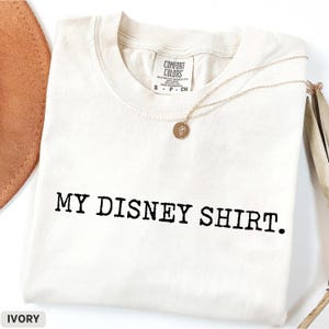 May include: Ivory-colored t-shirt with the text "MY DISNEY SHIRT." printed in black. A gold necklace with a small pendant rests on the shirt. The shirt is folded and displayed on a white surface.
