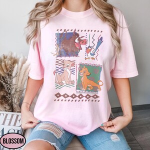 May include: A light pink t-shirt featuring a colorful graphic of characters from The Lion King. The design includes images of Pumbaa, Timon, Simba, and Nala, set against a geometric background. The shirt has short sleeves and a crew neck.