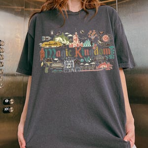 May include: Dark gray t-shirt with a colorful graphic of a theme park, including a castle, clock, and the words "Magic Kingdom." The design features various illustrations and is printed on a short-sleeved, oversized tee.
