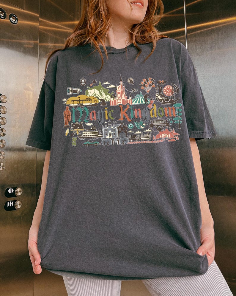 May include: Dark gray t-shirt with a colorful graphic of a theme park, including a castle, clock, and the words "Magic Kingdom." The design features various illustrations and is printed on a short-sleeved, oversized tee.