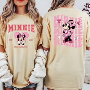 May include: Cream-colored t-shirt featuring a pink graphic of Minnie Mouse. The front design includes the word "MINNIE" in a pink collegiate font, with "EST. 1928" and "MINNIE MOUSE" below. The back displays a larger Minnie Mouse graphic and the word "MINNIE" repeated.