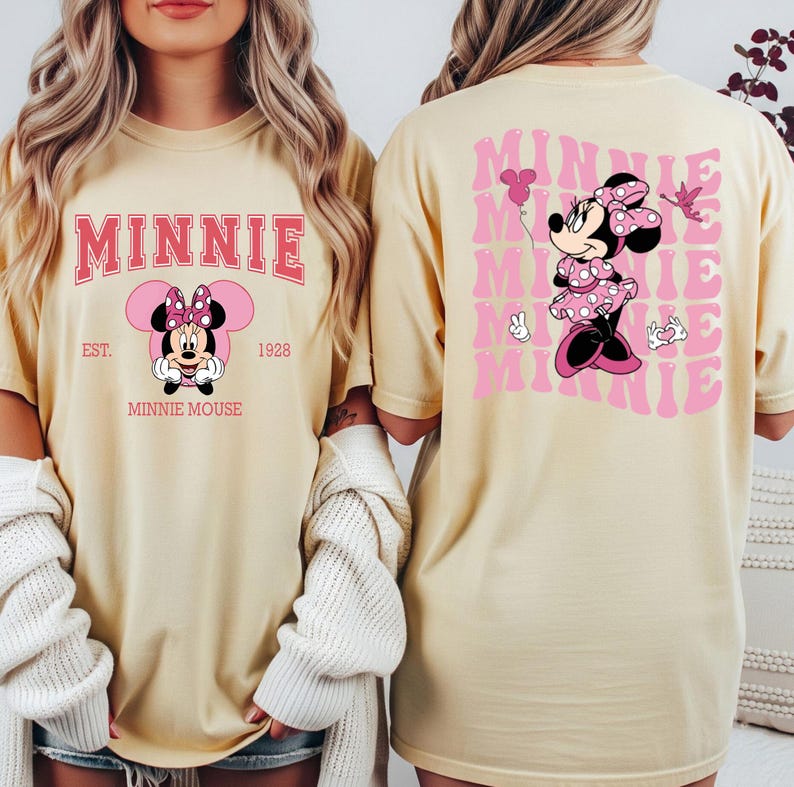 May include: Cream-colored t-shirt featuring a pink graphic of Minnie Mouse. The front design includes the word "MINNIE" in a pink collegiate font, with "EST. 1928" and "MINNIE MOUSE" below. The back displays a larger Minnie Mouse graphic and the word "MINNIE" repeated.