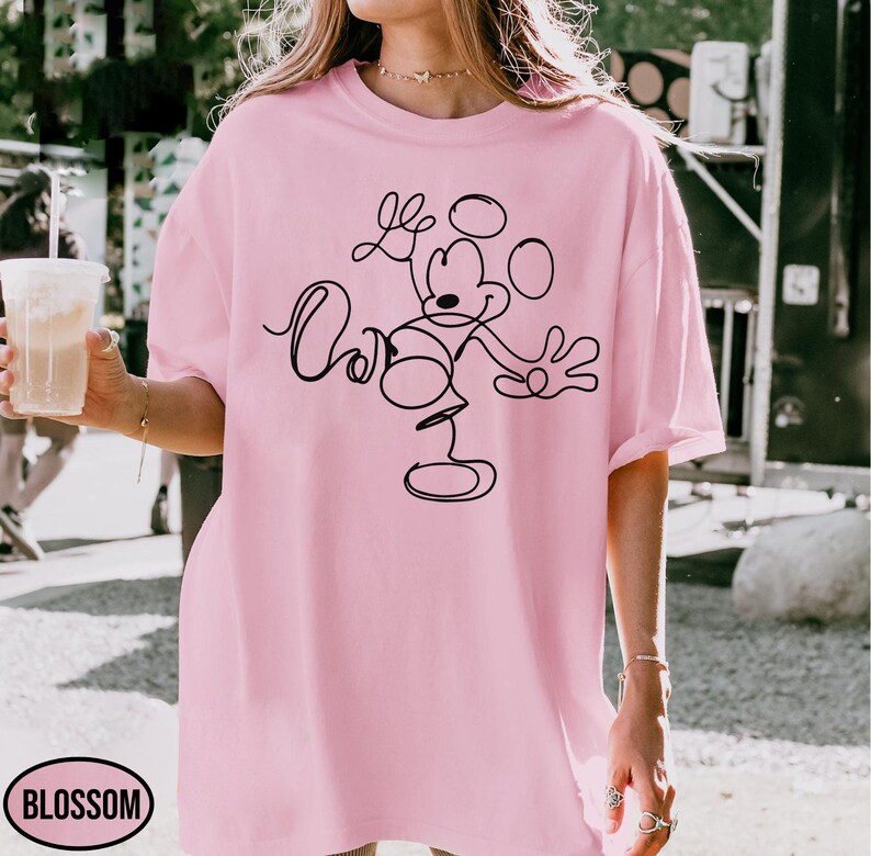 May include: A light pink t-shirt featuring a black line drawing of Mickey Mouse. The design is a continuous line, depicting Mickey in a playful pose. The shirt has short sleeves and a relaxed fit, perfect for casual wear.