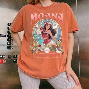 May include: Orange t-shirt with the word "MOANA" in a pink, stylized font. The shirt features a circular graphic of a woman in a floral crown, surrounded by flowers and tropical scenery. The shirt is a short-sleeved, crew neck style.