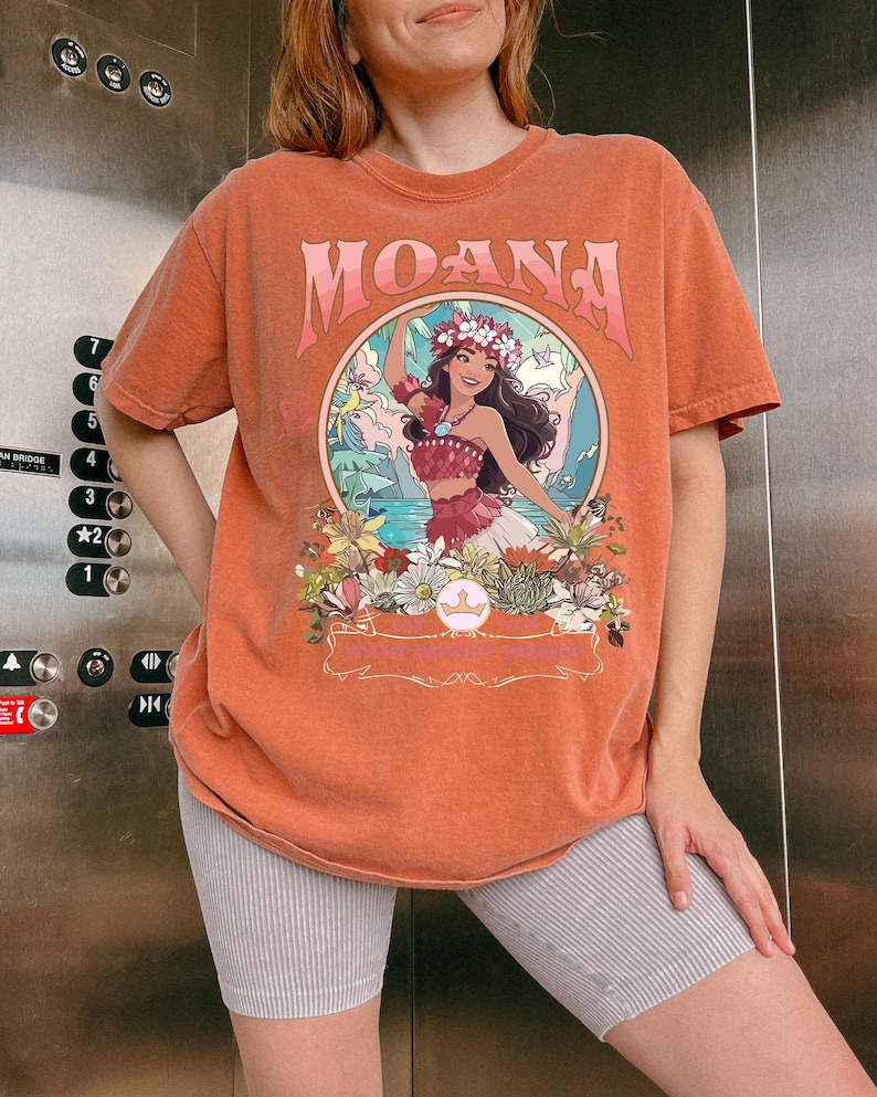 May include: Orange t-shirt with the word "MOANA" in a pink, stylized font. The shirt features a circular graphic of a woman in a floral crown, surrounded by flowers and tropical scenery. The shirt is a short-sleeved, crew neck style.
