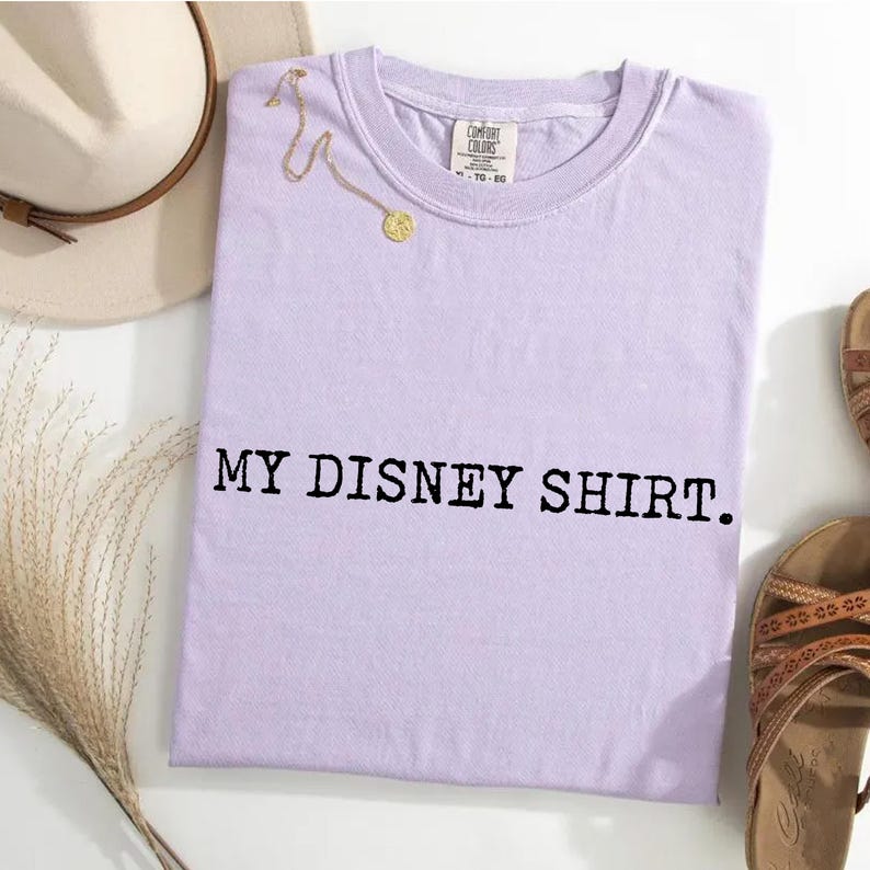 May include: A light purple t-shirt with the text "MY DISNEY SHIRT." A gold necklace and a beige hat are next to the shirt. The shirt is folded on a white surface.