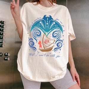 May include: Cream-colored t-shirt featuring a blue and teal graphic of a boat with a character, surrounded by waves and a palm tree design. The text "There is nowhere you can go that I won't be with you..." is printed on the shirt.