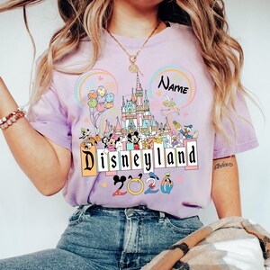 Op de afbeelding: Lichtpaars T-shirt met een kleurrijke Disneyland-afbeelding. Het ontwerp bevat een kasteel, verschillende Disney-personages, ballonnen en de tekst "Disneyland 2020". Er is ook ruimte voor een naam.