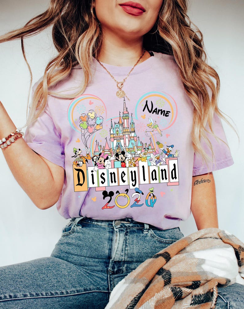 Op de afbeelding: Lichtpaars T-shirt met een kleurrijke Disneyland-afbeelding. Het ontwerp bevat een kasteel, verschillende Disney-personages, ballonnen en de tekst "Disneyland 2020". Er is ook ruimte voor een naam.