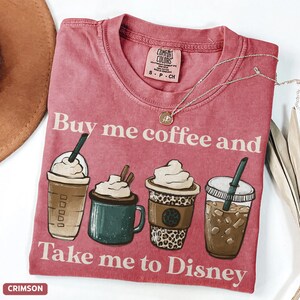 Puede incluir: Camiseta de color carmes&iacute; con el texto "Buy me coffee and Take me to Disney". La camiseta presenta ilustraciones de cuatro bebidas de caf&eacute; diferentes. Tambi&eacute;n es visible un collar plateado con un colgante.
