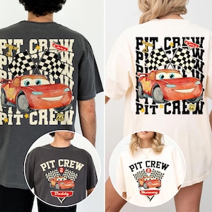 May include: Two t-shirts with a red race car graphic and the words "PIT CREW". One is dark grey, the other cream. The design includes checkered flags and the car's number. The shirts are likely for a family.