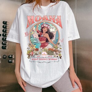 May include: White t-shirt featuring the word "MOANA" in pink lettering above a circular graphic. The graphic depicts a woman in a floral crown and traditional attire, surrounded by flowers and butterflies. The text "WALT DISNEY WORLD" is below the graphic.